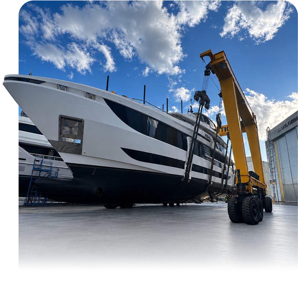 Large yacht beside a yellow mobile crane in a shipyard under a cloudy blue sky